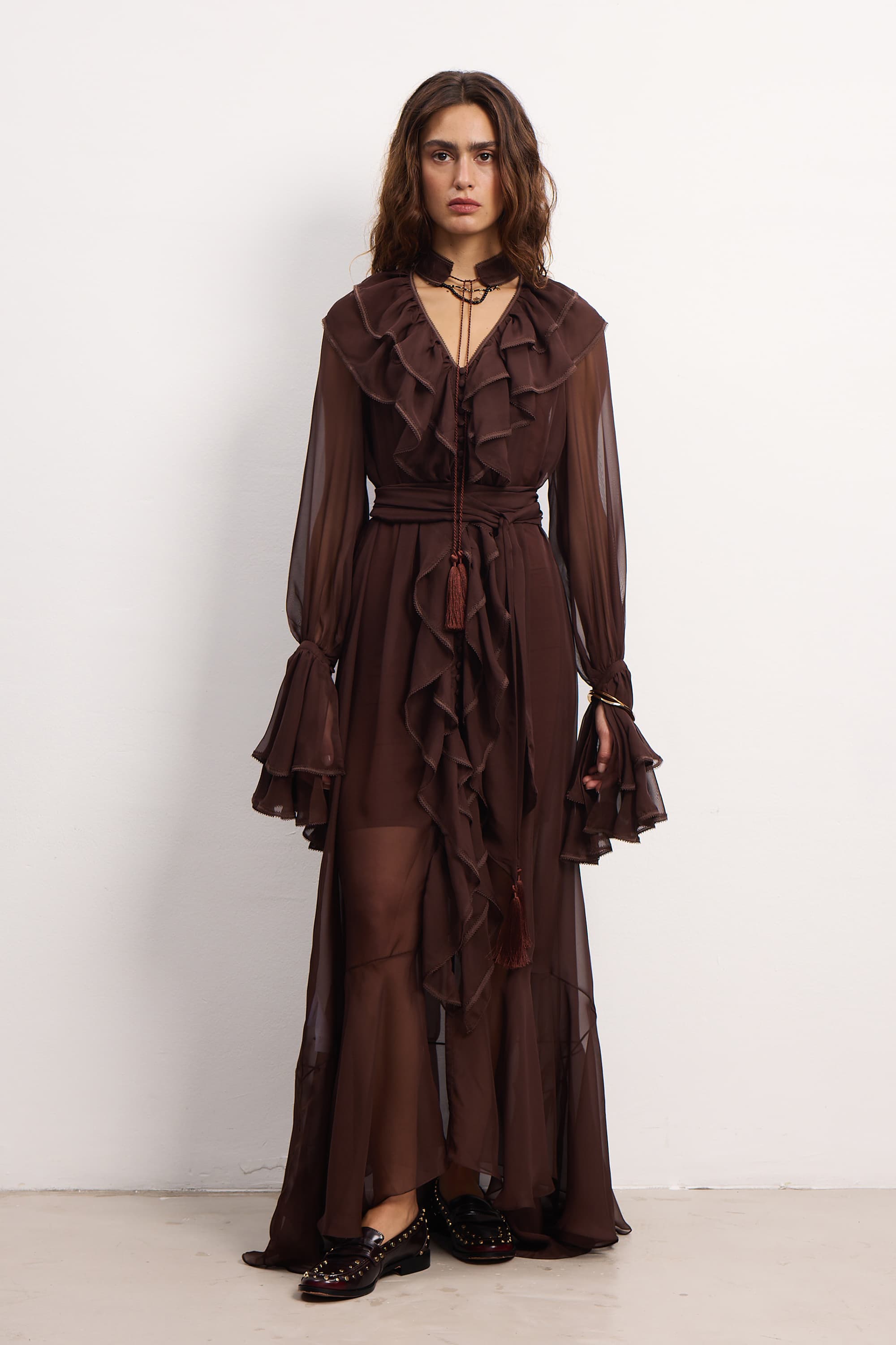 Model wearing a floor-length, sheer dark brown dress with cascading ruffles along the front, tiered sleeves, and a tassel choker