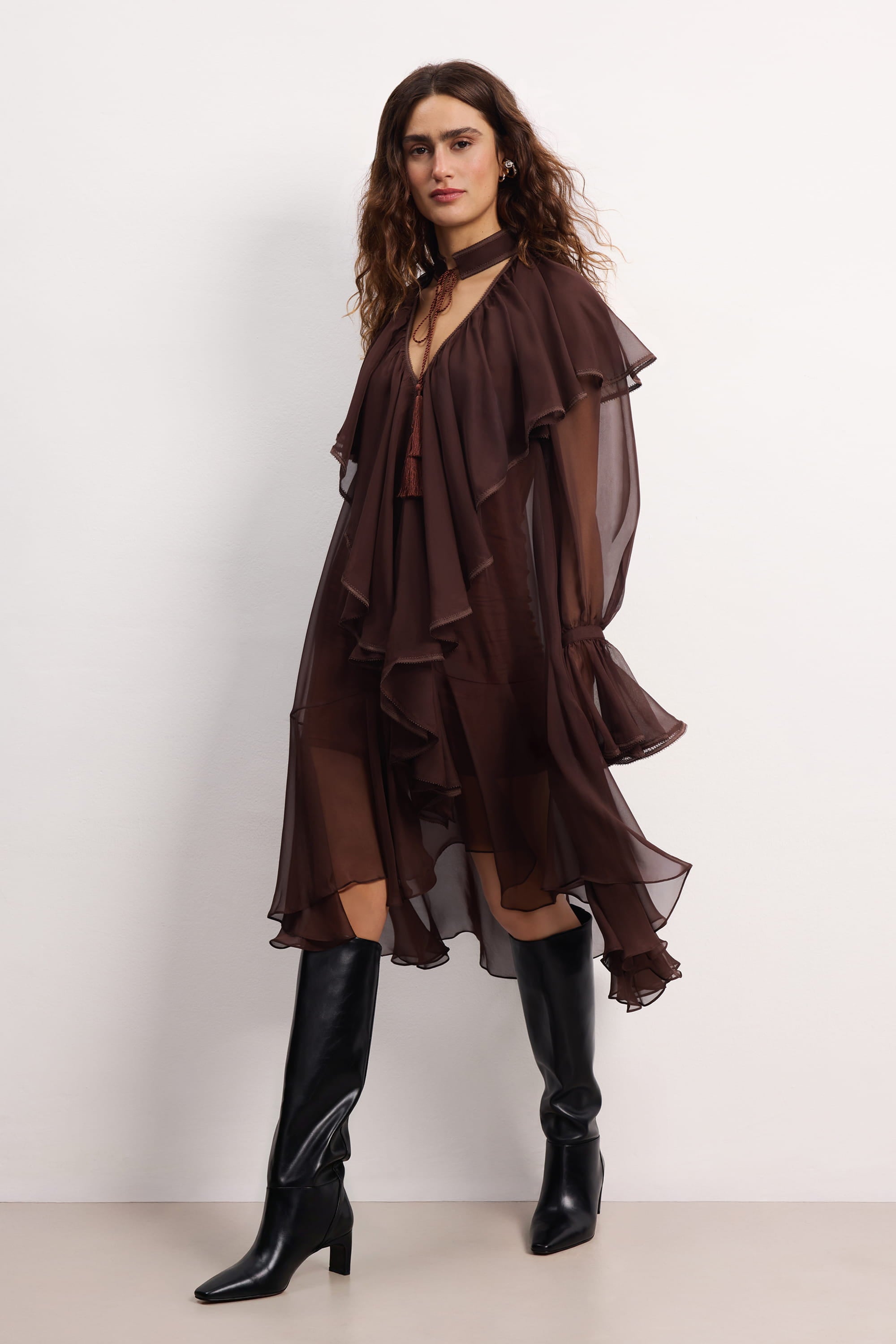 Woman wearing a brown ruffled Ocean Of Tenderness midi dress with black knee-high boots against a white background, side view