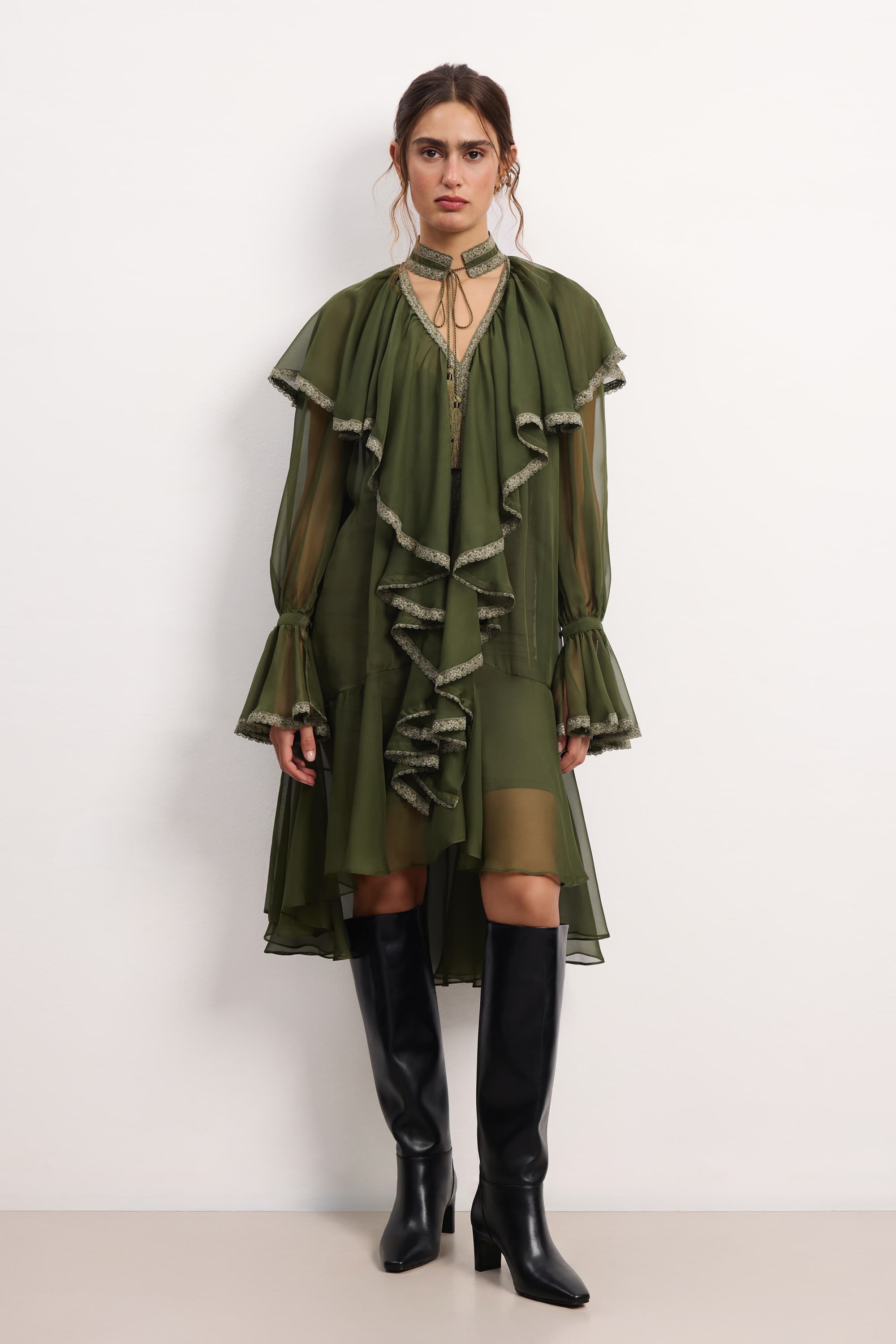 Woman wearing a olive ruffled Ocean Of Tenderness midi dress with black knee-high boots against a white background, front view