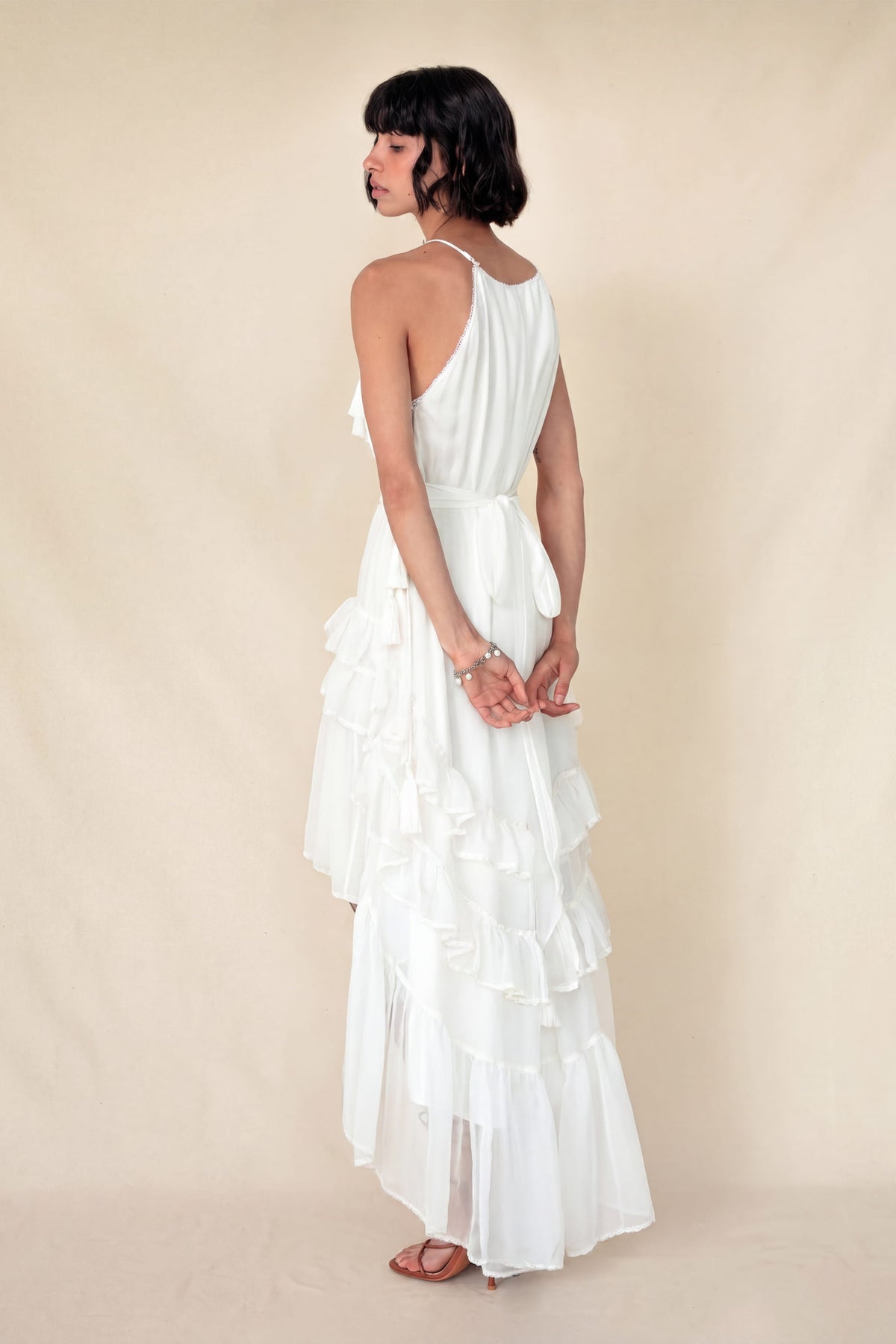 A woman wearing a white sleeveless maxi dress with a pleated bodice and tiered ruffle skirt, shown from the back against a plain beige studio backdrop.