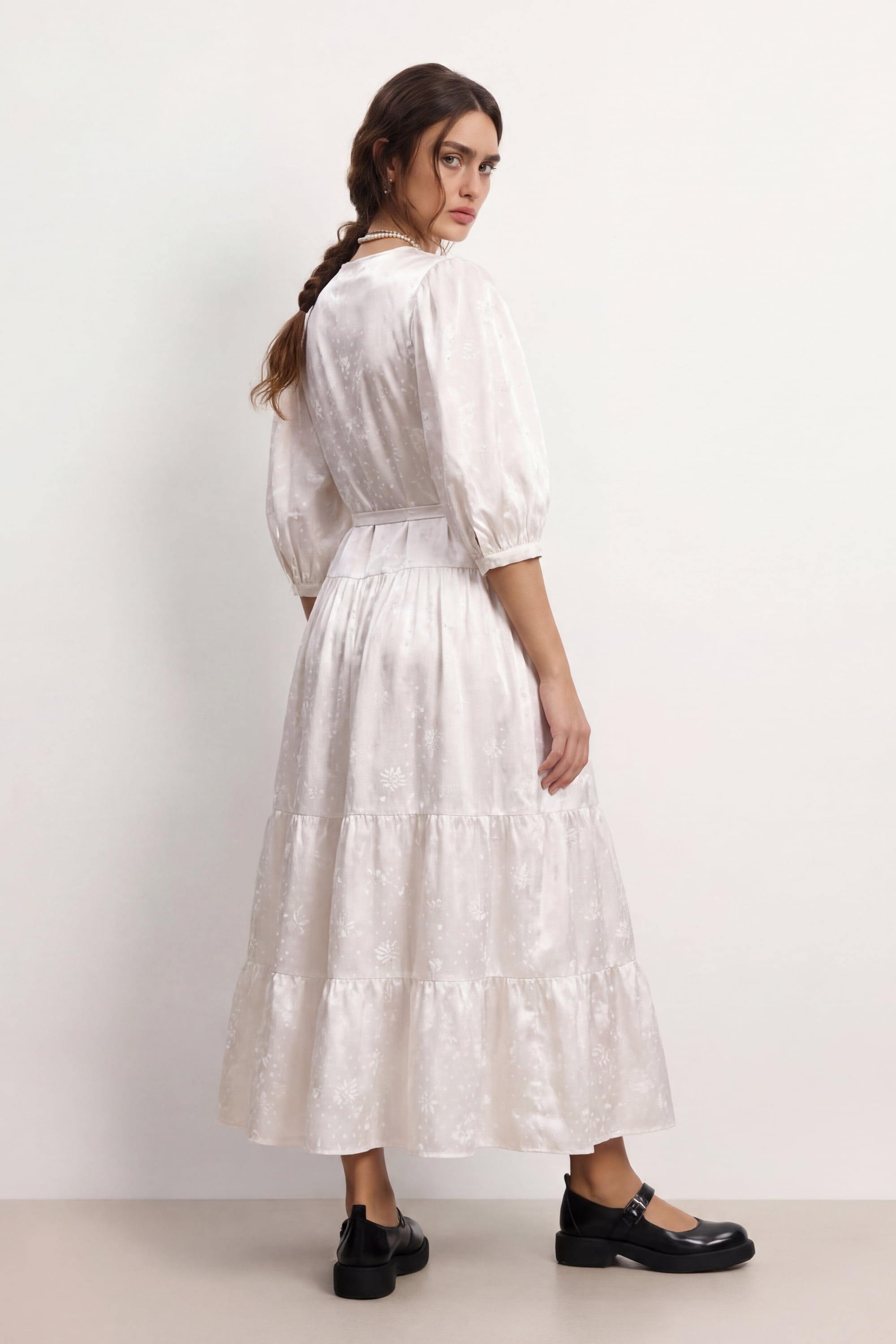 Woman wearing a soft white Haven dress with long sleeves and a belt, standing against a plain background, back view
