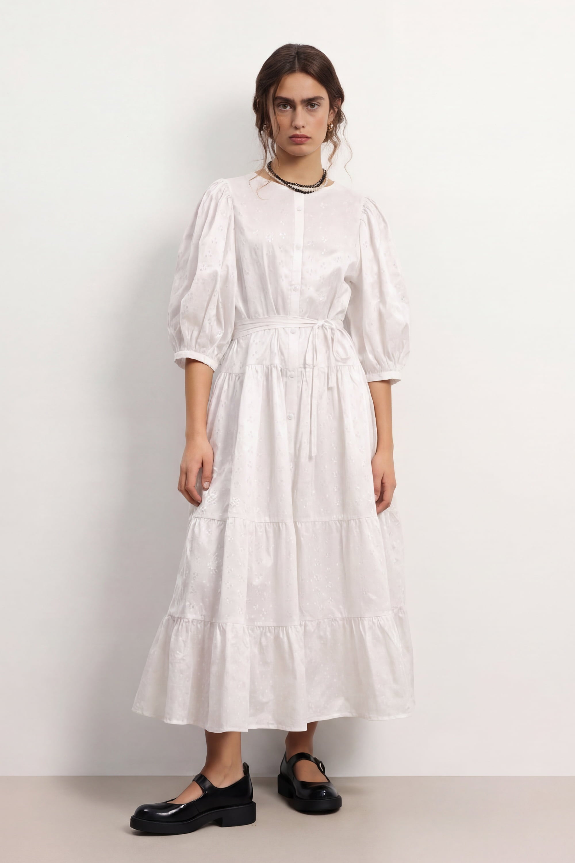 Woman wearing a soft white Haven dress with long sleeves and a belt, standing against a plain background, front view