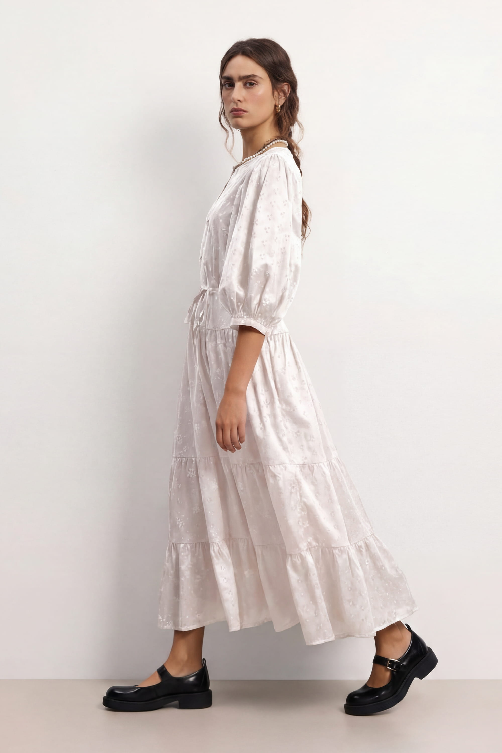 Woman wearing a soft white Haven dress with long sleeves and a belt, standing against a plain background, side view