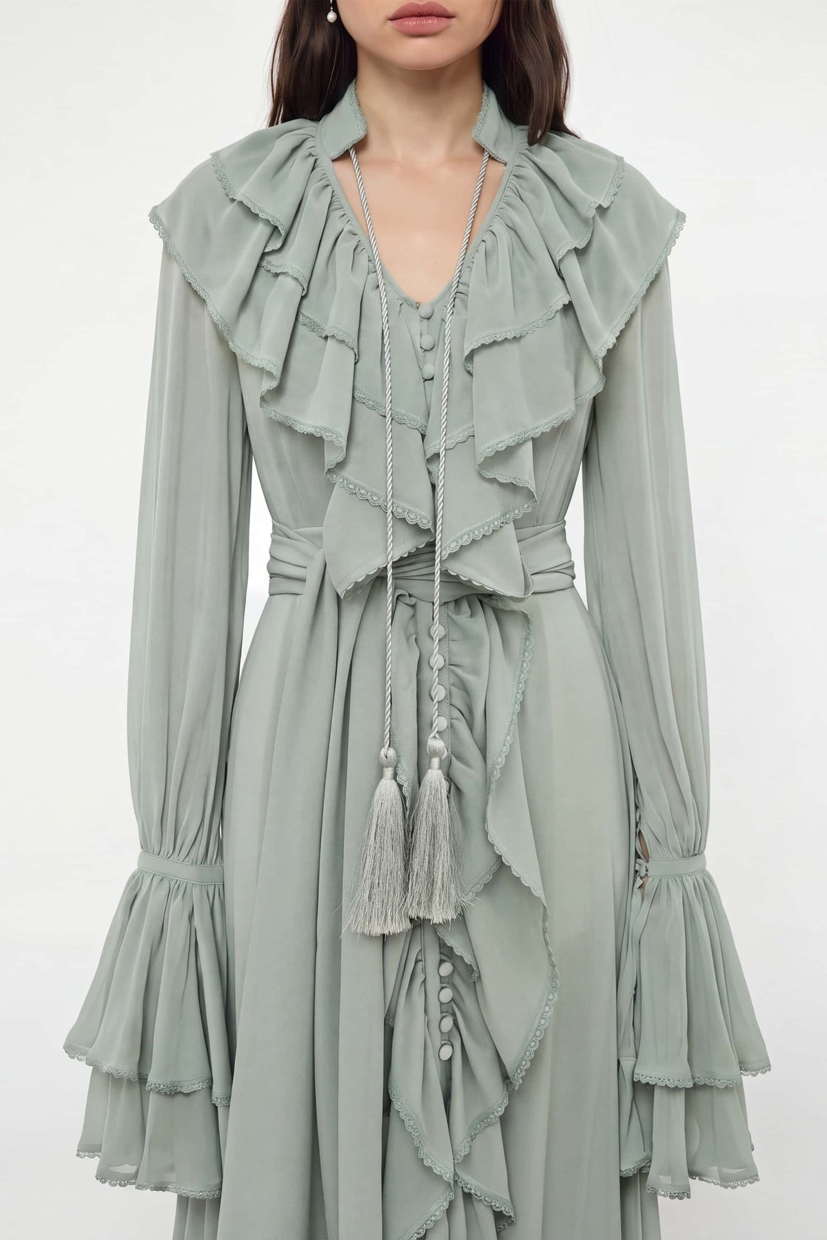 A woman wearing a sage green long-sleeve Ocean Of Tenderness dress with dramatic layered ruffles, a tie-neck detail, and tassel accents, photographed against a plain light background