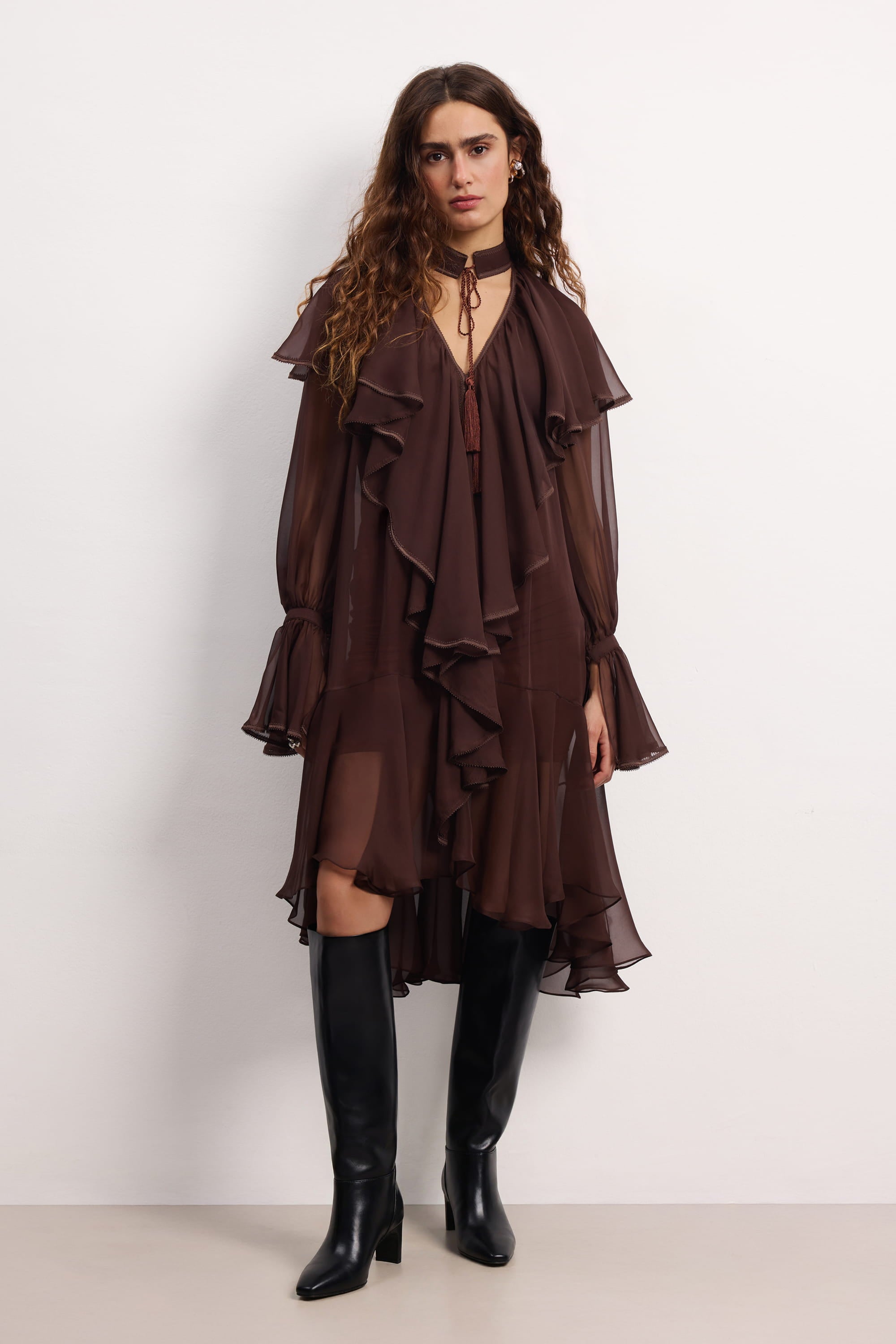 Woman wearing a brown ruffled Ocean Of Tenderness midi dress with black knee-high boots against a white background, front view