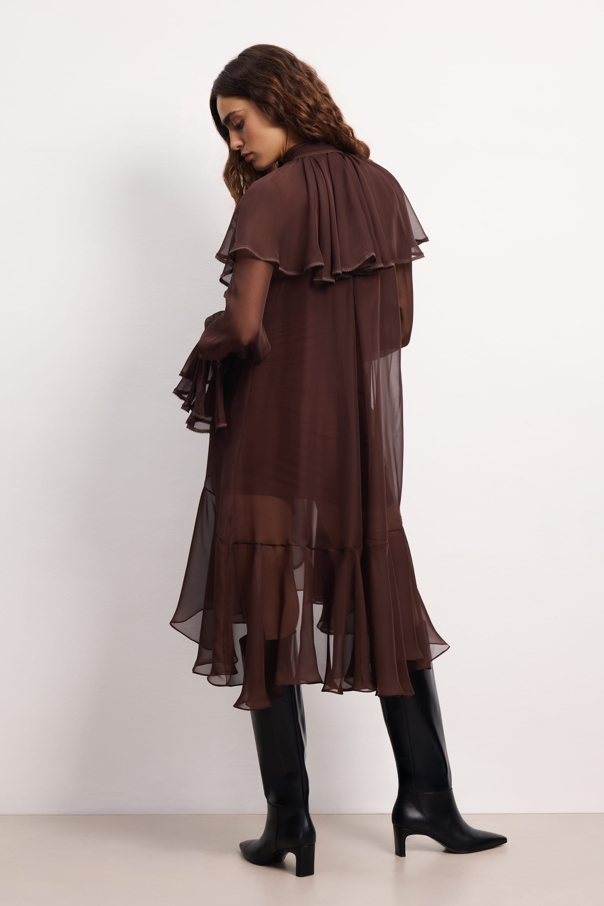 Woman wearing a brown ruffled Ocean Of Tenderness midi dress with black knee-high boots against a white background, back view