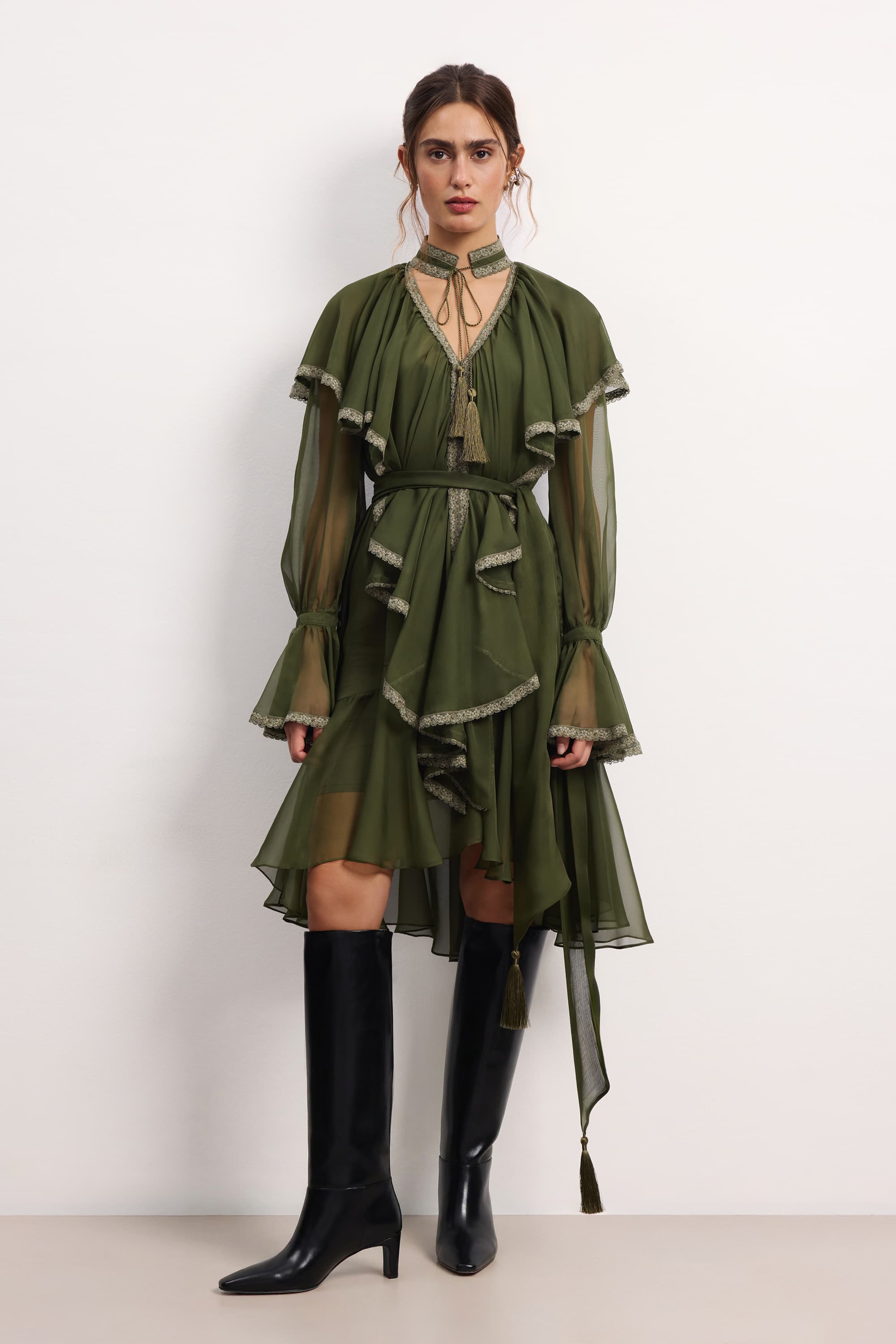 Woman wearing a olive ruffled Ocean Of Tenderness midi dress with black knee-high boots against a white background, front view