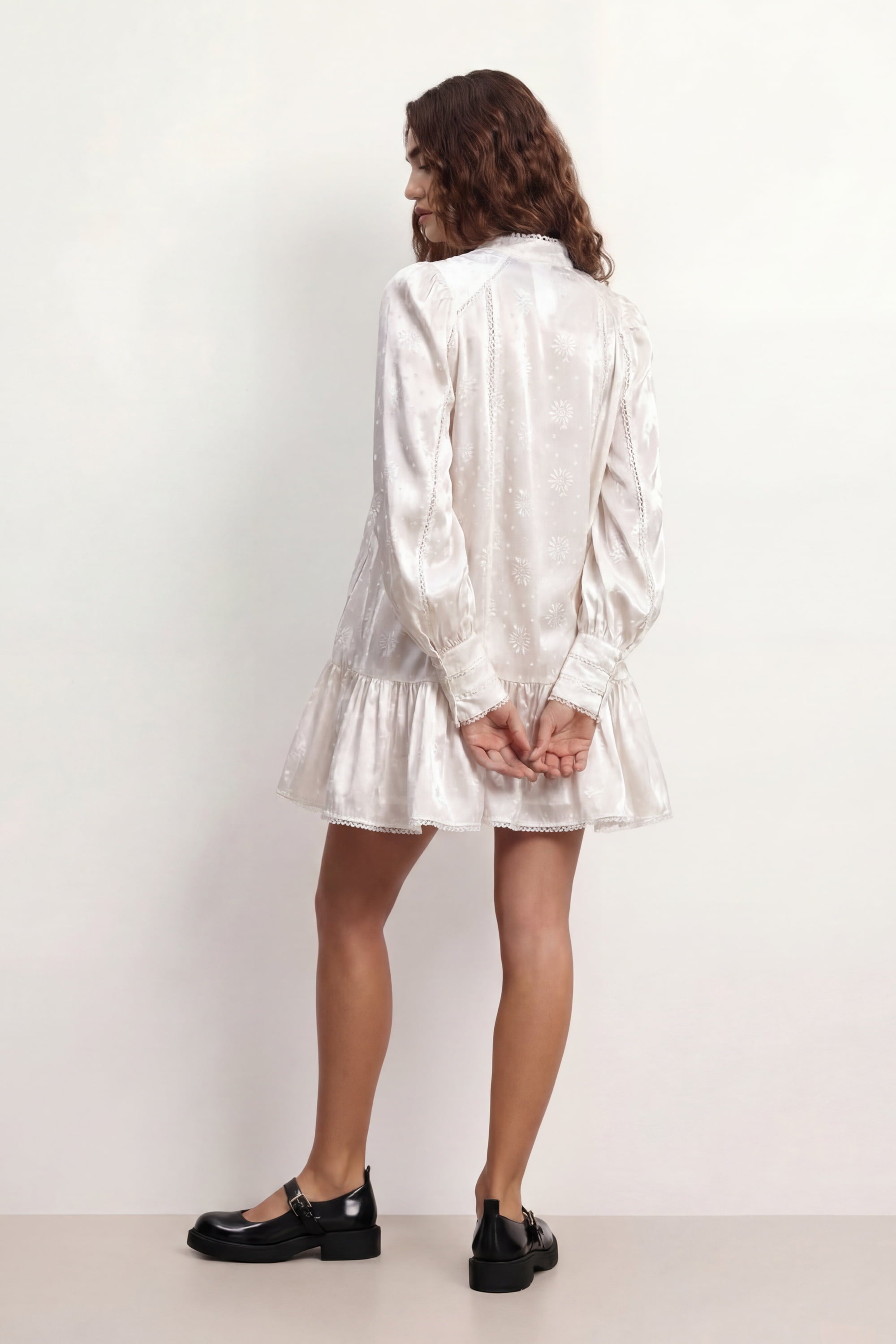 Woman wearing a soft white Wildflower dress with long sleeves and lace details against a plain background, back view