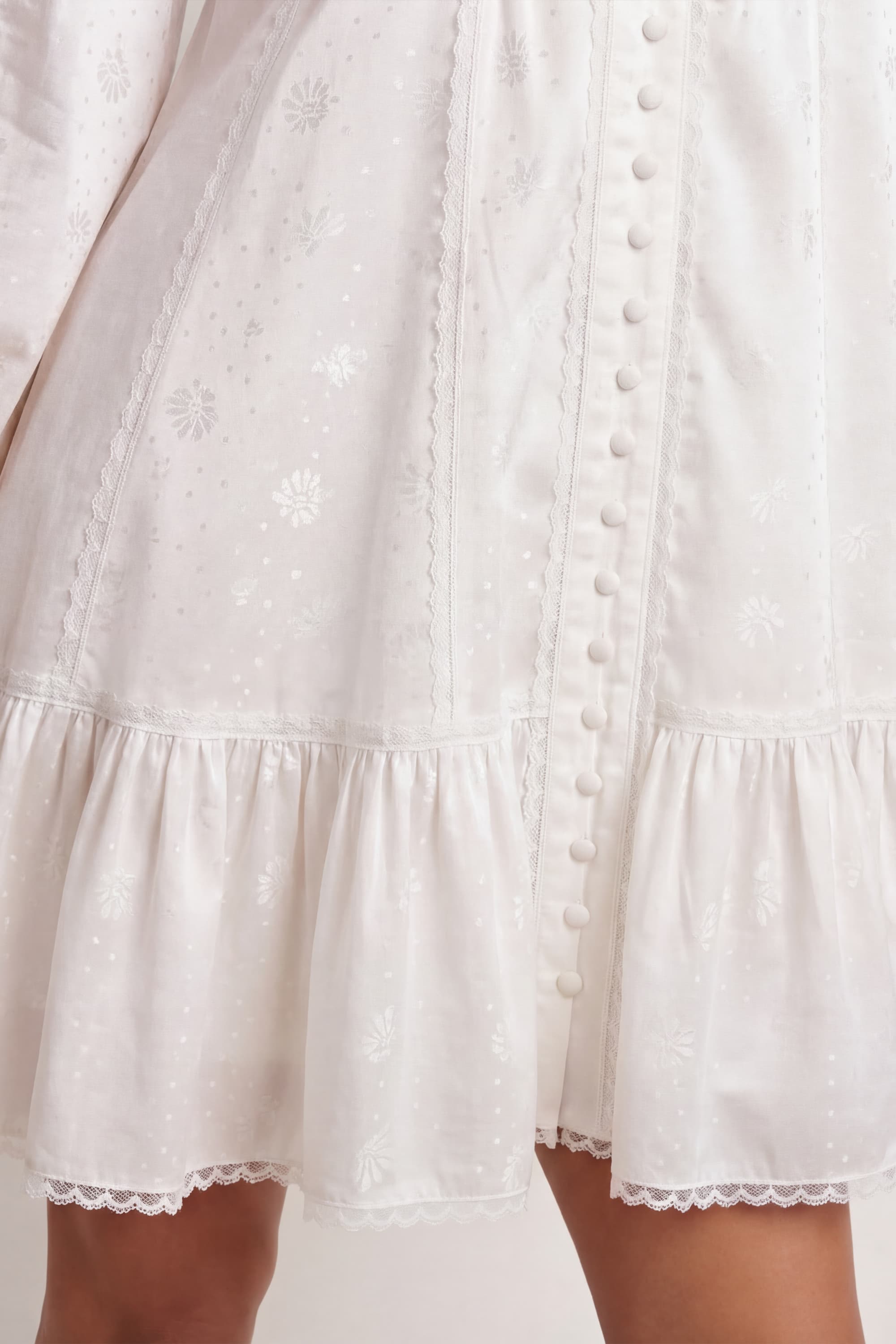 Woman wearing a soft white Wildflower dress with long sleeves and lace details against a plain background, details front view